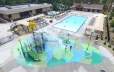 In the foreground the splash pad area is shown with children playing on the equipment. Behind the Splash Pad the zero entry pool is shown with people swimming in it and lounging in the chairs around it. In the far left of the photo the hot tub area is shown.