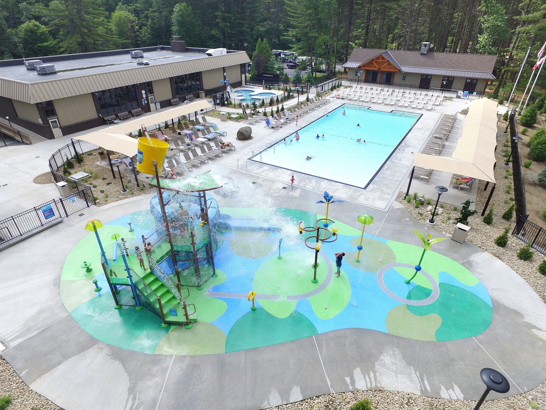 In the foreground the splash pad area is shown with children playing on the equipment. Behind the Splash Pad the zero entry pool is shown with people swimming in it and lounging in the chairs around it. In the far left of the photo the hot tub area is shown.