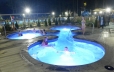 In the foreground the hot tub area is shown at night with guests lounging in it.   In the background the dump bucket and slide of the splash pad area is shown.