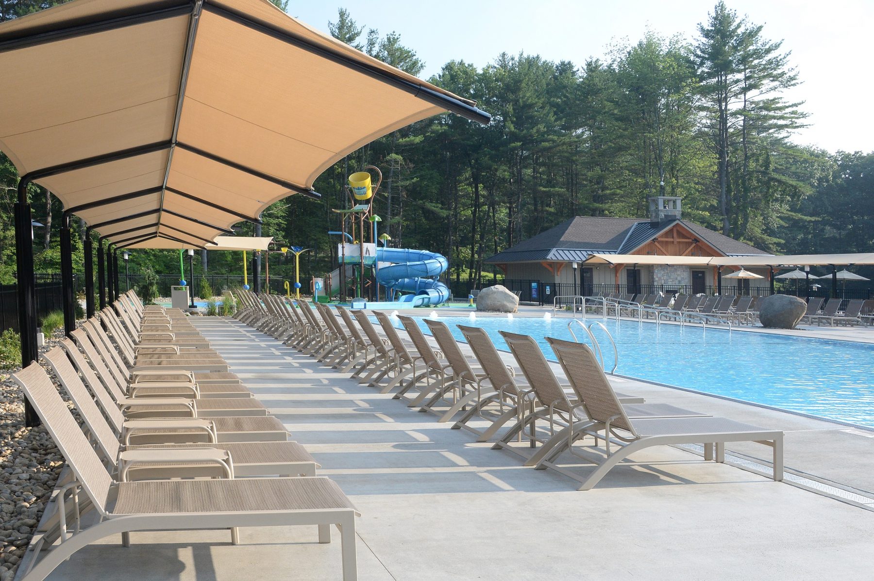 In the foreground the chairs are shown in two rows.  The back row is under a large shade and the front row is out in the sun. In front of the chairs the pool is shown.  In the background of the photo the splashpad area including the slide and dump bucket are shown. In the far right corner the Cascade Cove Bathroom facilities are shown.