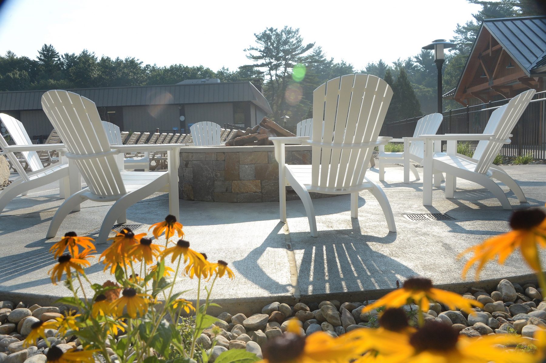 In this photo there is adirondack chairs surrounding a stone fire pit. Wood is stacked in the fire pit for the fire. Surrounding this area there are stones and flowers.