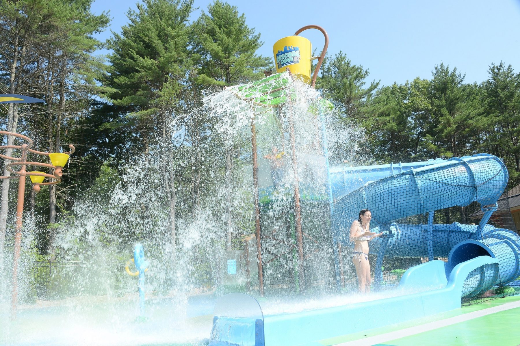 In the foreground of this photo the bottom of the slide is shown. Behind the slide there is a young woman being sprayed by the various sprayers.  The dump bucket is shown upright at the top of the photo in the center. To the right side of the photo the enclosed slide is shown.