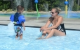 A woman is sitting in the entry area of the pool facing a young little boy who is standing in the entry of the pool. In the background some of the sprayers on the splash pad are shown.