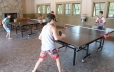 In the foreground of this photo both ping pong tables are shown. They are both being occupied by young boys playing ping pong.  In the background of the photo a young woman is shown sitting in one of the chairs watching the boys play.