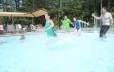 In the foreground of the photo a woman is shown running into the zero entry pool.  In the background of the photo a man accompanied by a young boy and a young girl are shown running into the pool. To the far left of the photo guests are shown lounging in the shaded area on the pool deck.
