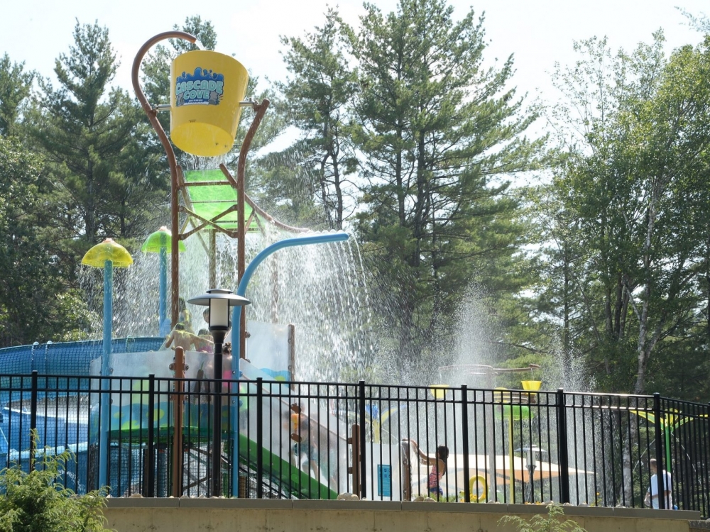 Splash pad with blue twisting slide, spray features, large yellow dump bucket, multiple children climbing steps up to slide, surrounded by black fence