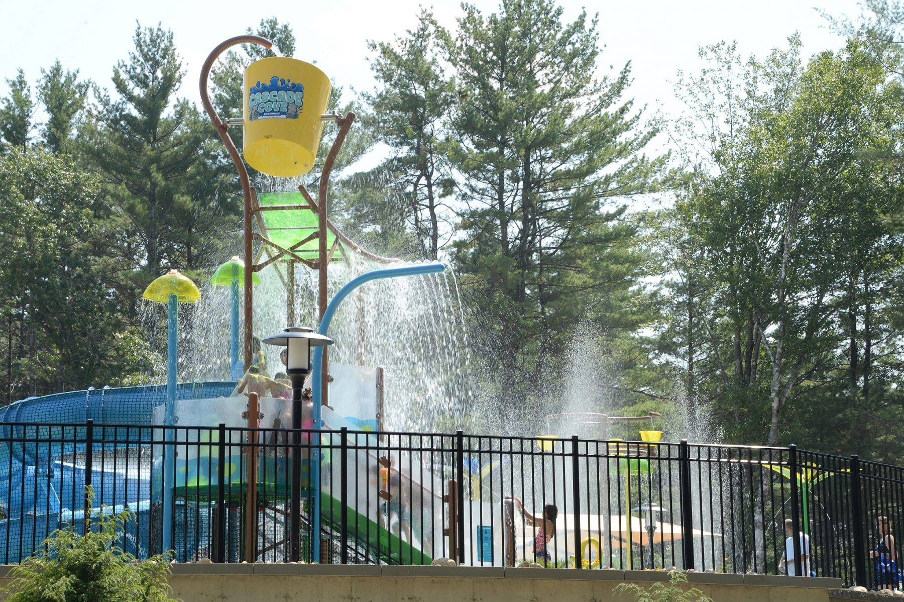 Splash pad with blue twisting slide, spray features, large yellow dump bucket, multiple children climbing steps up to slide, surrounded by black fence