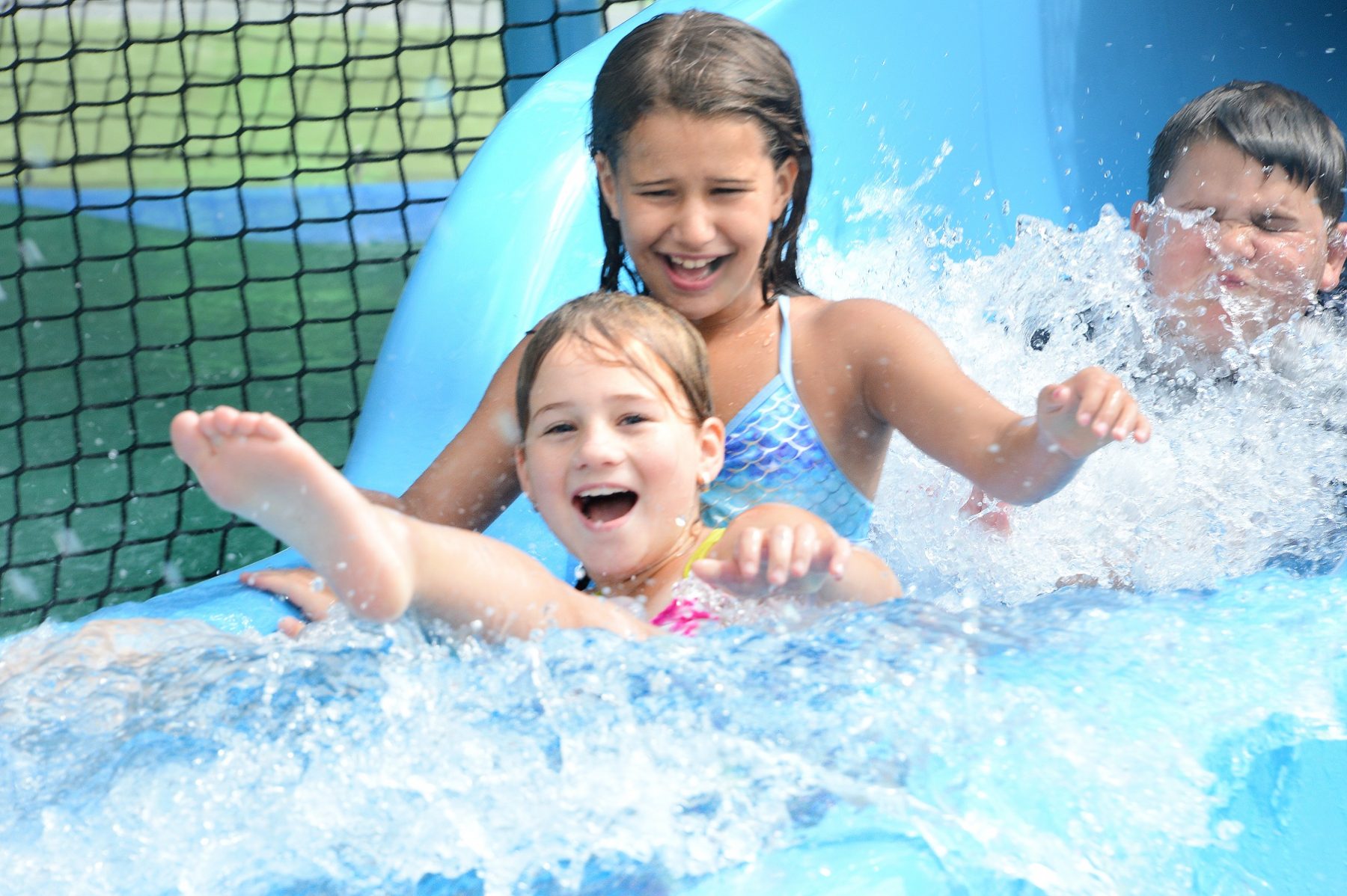 In the foreground of the photo two young girls are seen sliding down to the bottom of the slide laughing and smiling.  Behind them is a young boy being splashed by the girls in front of him.