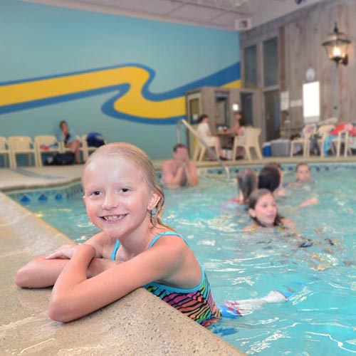 Child inside the indoor pool, holding onto the edge and smiling. In the background are other guests swimming