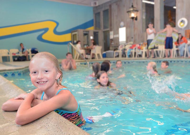 Child inside the indoor pool, holding onto the edge and smiling. In the background are other guests swimming