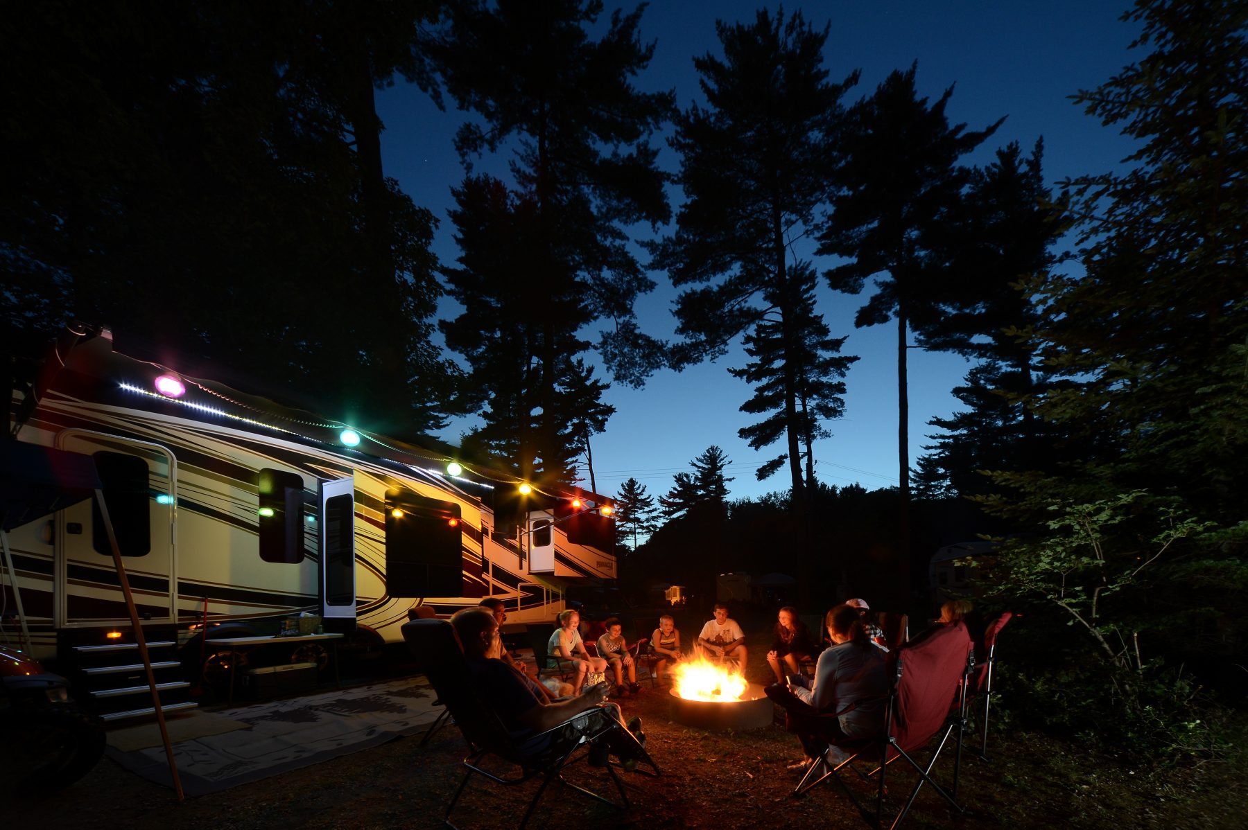 family sitting by campfire in front of RV at sunset under trees