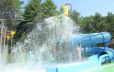 In the foreground of this photo the bottom of the slide is shown. Behind the slide there is a young woman being sprayed by the various sprayers.  The dump bucket is shown upright at the top of the photo in the center. To the right side of the photo the enclosed slide is shown.