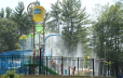 Splash pad with blue twisting slide, spray features, large yellow dump bucket, multiple children climbing steps up to slide, surrounded by black fencee twisting slide, spray features, large yellow dump bucket, multiple children climbing steps up to slide, surrounded by black fence