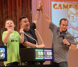 Game show madness night at the French Mountain Playhouse. The host to the right is high fiving a gentleman who is wearing a dark grey shirt and smiling. Next to him is a younger boy in a lime green t shirt smiling and giving two thumbs up! In front of them are microphones, and their score boards which shows 50 points each in green and purple lights. To the left is another score board showing 25 points.