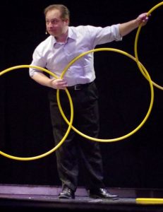 Illusionist David Garrity with 3 connected hoola hoops on stage with a head set microphone