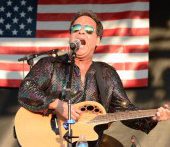 Man playing guitar singing into a microphone with an American Flag draped behind him.